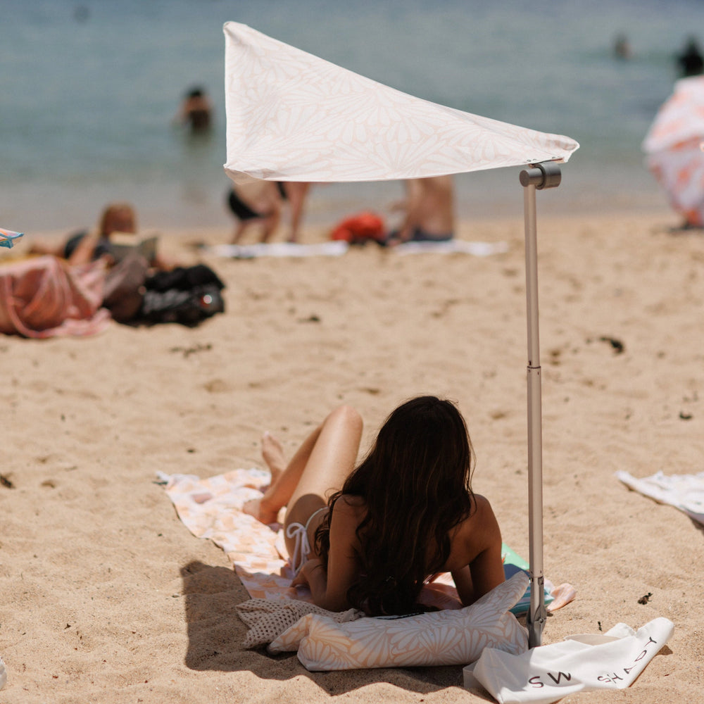 Woman relaxing on the sand under the cream Soothe Swim Shady beach canopy with a warm petal pattern